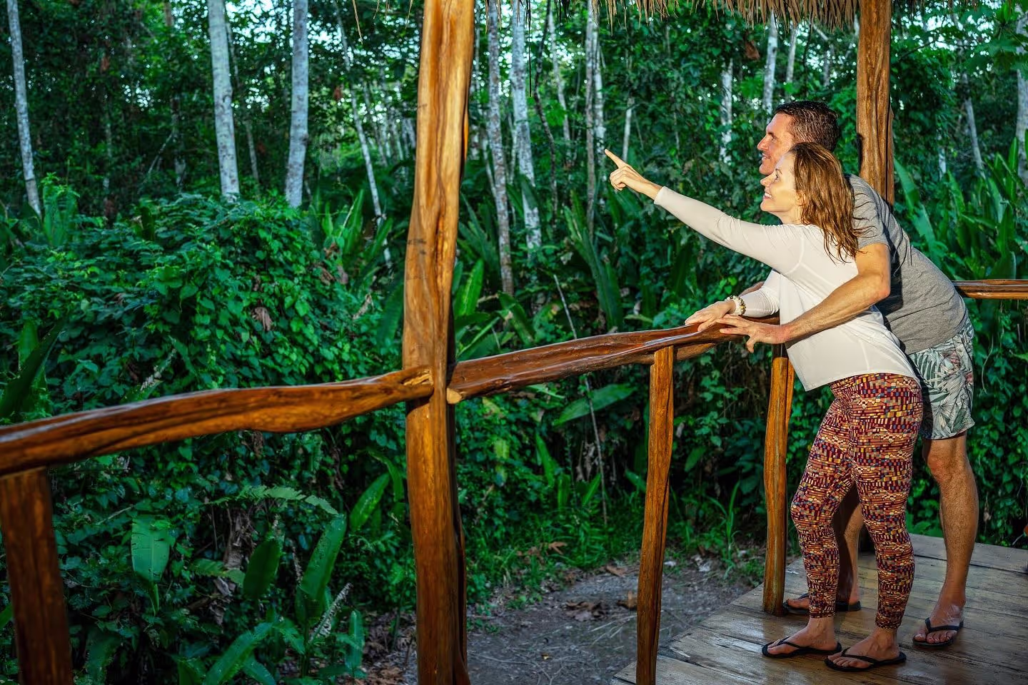 A man and woman lean on a rustic wooden railing, with the woman pointing excitedly toward dense green jungle foliage in the Amazon rainforest at an outdoor retreat.