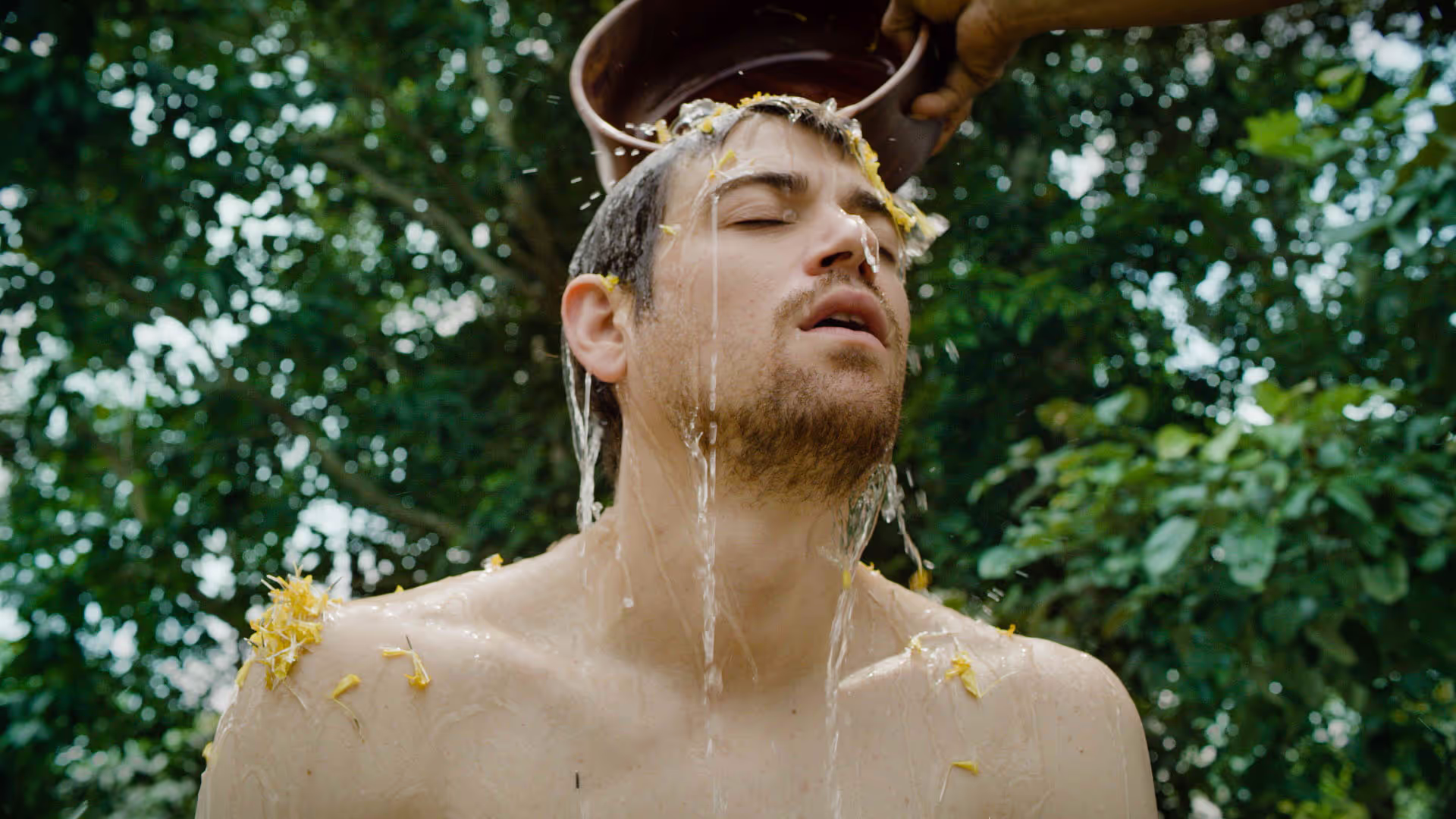 A man with eyes closed, immersed in a cleansing ritual as water mixed with yellow flower petals is poured over his head from a wooden bowl. Surrounded by dense jungle greenery, the scene captures a moment of spiritual purification and connection with nature.