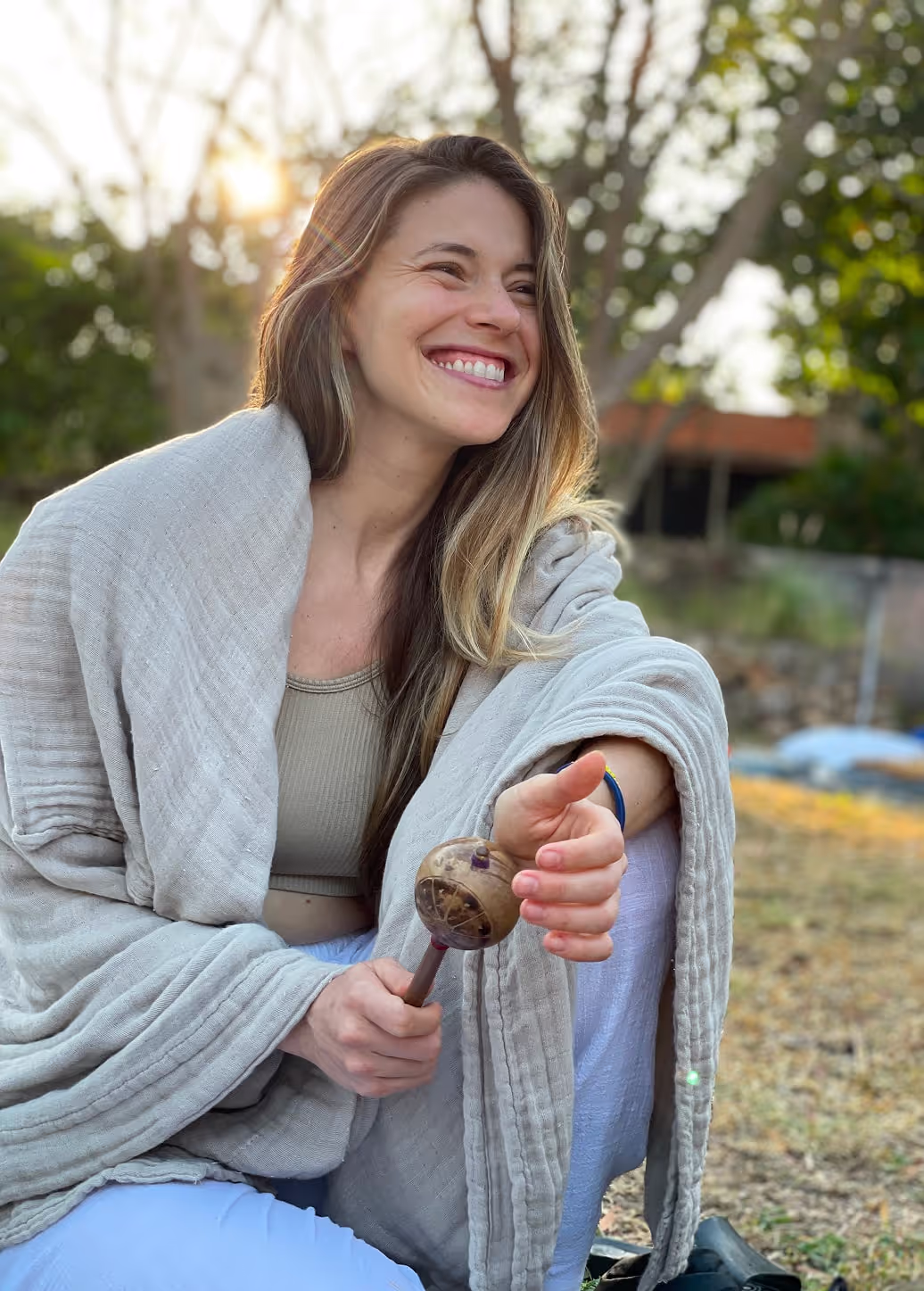 A joyful woman wrapped in a light beige shawl, holding a traditional shamanic rattle in her right hand. She is seated outdoors with sunlight filtering through trees in the background, creating a warm and peaceful atmosphere. Her expression is bright and genuine, capturing a moment of happiness and connection.