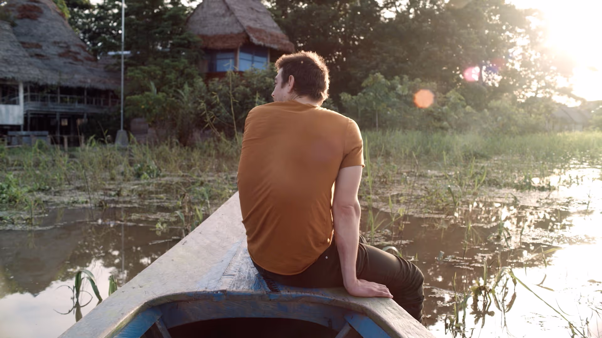 A man in a burnt orange shirt sits contemplatively on the edge of a wooden canoe, surrounded by shallow waters and lush greenery, with the warm glow of the setting sun illuminating the scene near an Amazon retreat center.