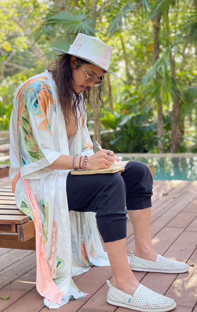 A man wearing a floral kimono and white hat sits on a wooden bench near a pool, deeply focused on writing in a notebook, surrounded by lush greenery and tranquil natural light.