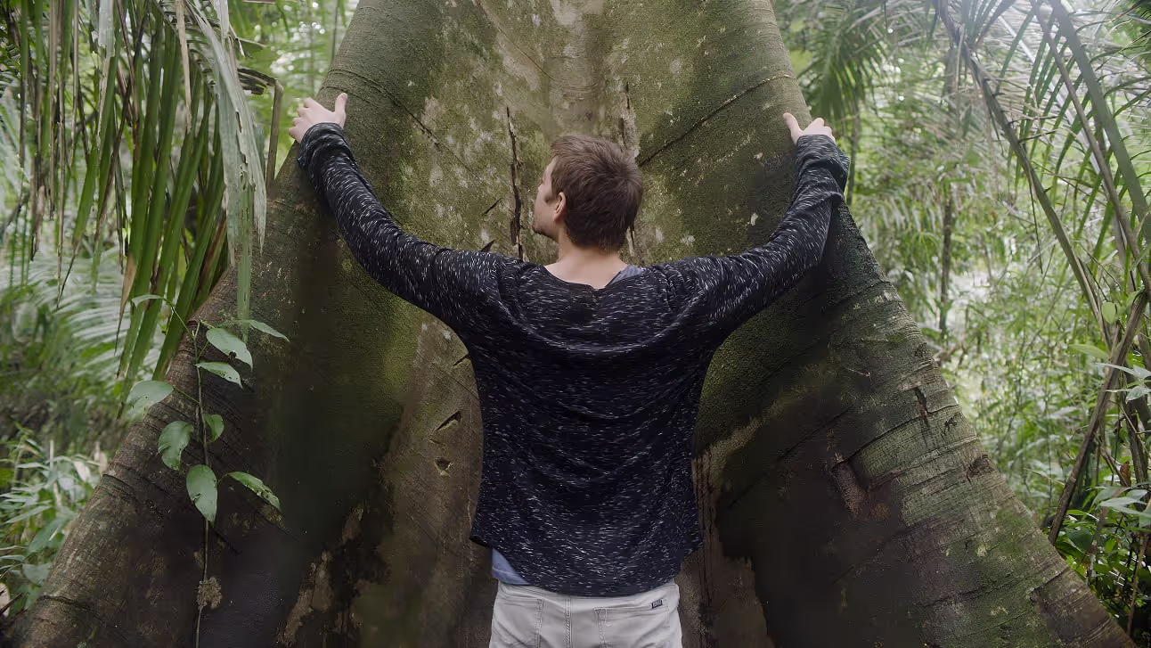 A man stands in a lush jungle with arms outstretched, holding onto the massive buttress roots of an ancient rainforest tree, symbolizing a deep connection to nature.