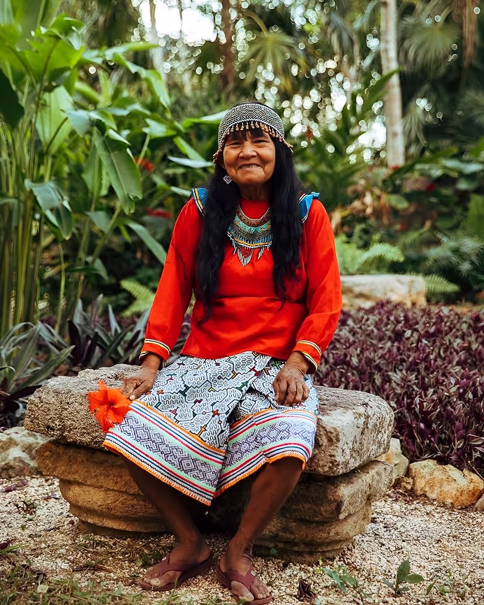 A warm and wise elder woman, dressed in traditional attire with vibrant patterns and beadwork, sits peacefully on a stone bench surrounded by tropical greenery and blooming plants. Her joyful expression radiates harmony with nature.