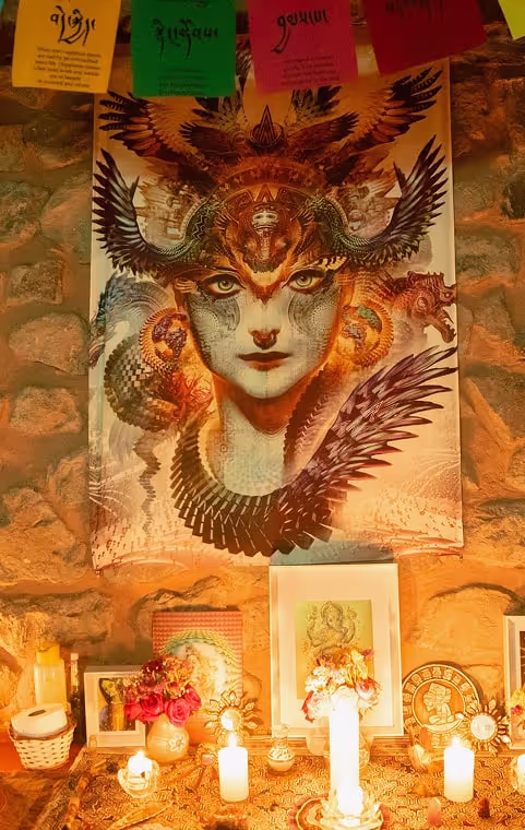 A spiritual altar adorned with candles, flowers, and sacred objects sits below a powerful visionary art tapestry depicting a mystical winged figure. Prayer flags hang above, enhancing the ceremonial ambiance.