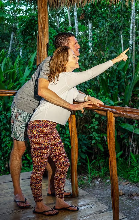 A couple stands joyfully on a wooden deck overlooking the rainforest, embraced and smiling as the woman points excitedly toward the treetops. Surrounded by lush greenery, the moment captures wonder, connection, and the tranquil magic of the jungle retreat experience.