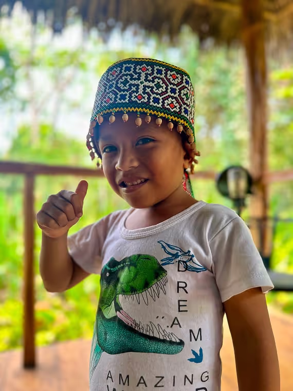Young child smiling and giving a thumbs up while wearing a traditional beaded hat and a dinosaur t-shirt, standing under a thatched roof in a lush jungle setting.