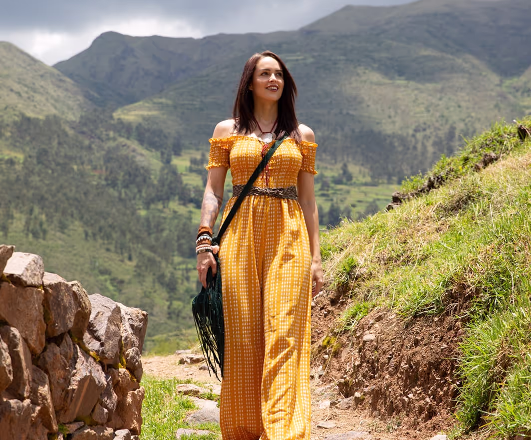 Woman in a bright yellow dress walking confidently along a scenic mountain path in the Sacred Valley, Peru—surrounded by lush greenery and ancient stone ruins.