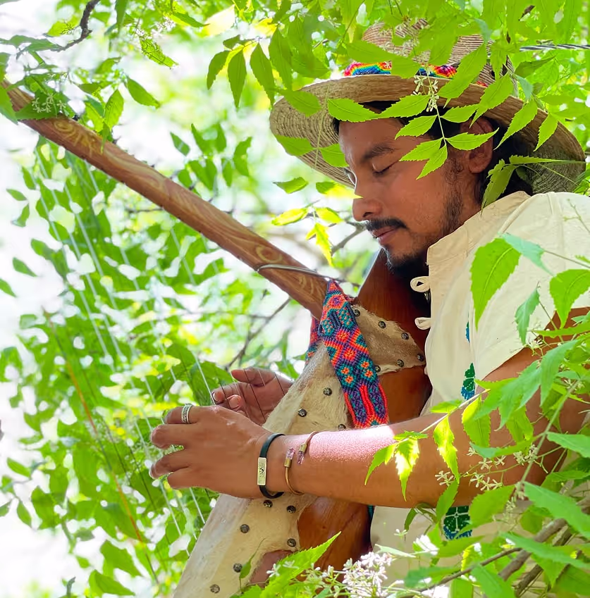 Shaman in traditional attire playing a handcrafted harp among lush green leaves, eyes closed in peaceful concentration during a sacred musical ceremony.