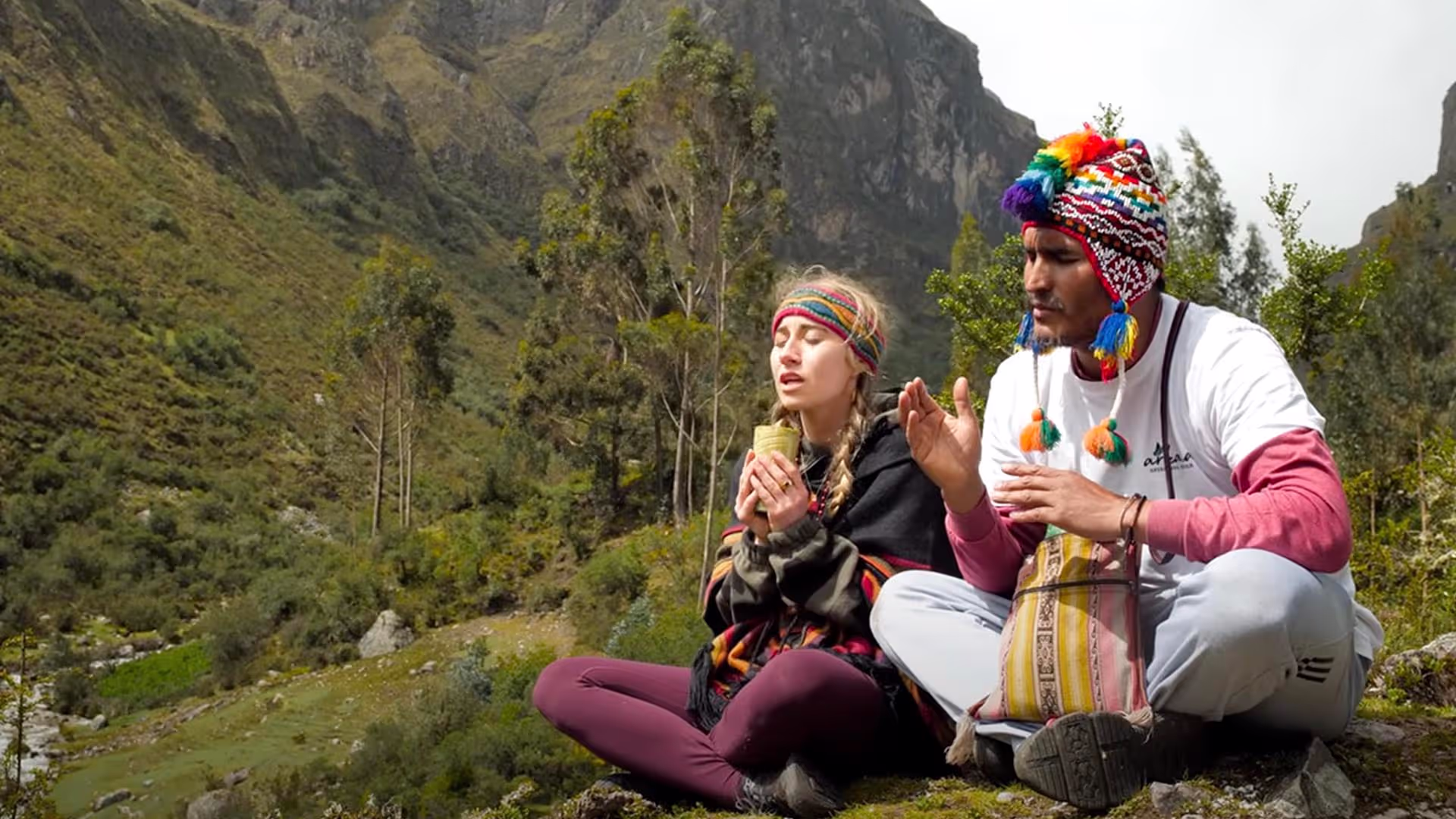 Shaman and participant sitting cross-legged in meditation during a mountain healing ceremony, holding a ceremonial cup and wearing traditional Andean attire in the Sacred Valley of Peru.