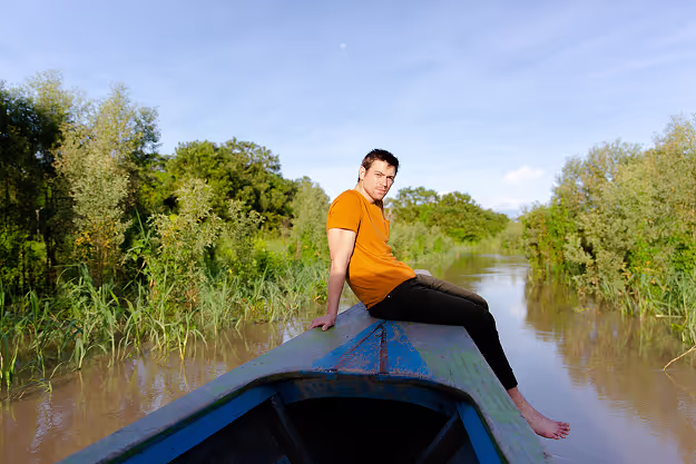 Man sitting barefoot on the front of a wooden boat as it moves through a narrow jungle river, surrounded by dense greenery under a bright blue sky.
