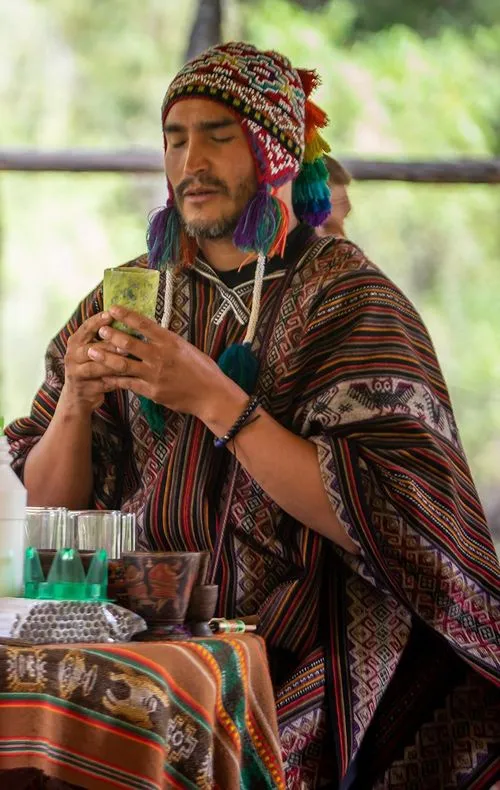 Shaman dressed in traditional Andean clothing holds a ceremonial cup with closed eyes, engaging in a prayer or blessing ritual during a pant medicine ceremony.