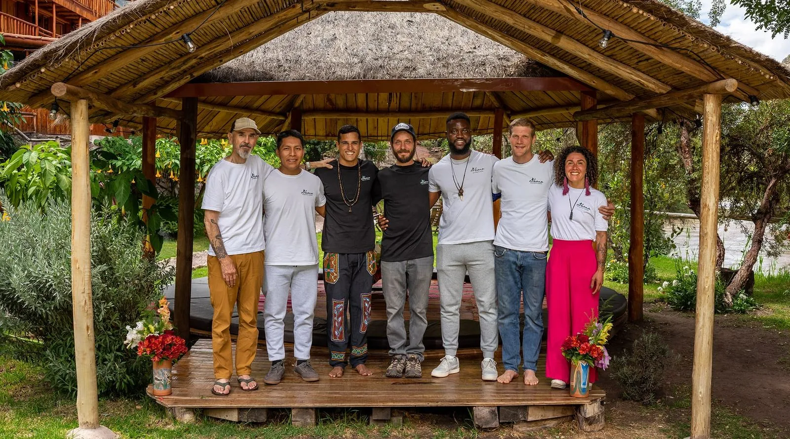 Group photo of diverse retreat staff standing together under a thatched-roof gazebo, smiling and united as part of the ayahuasca healing center team.