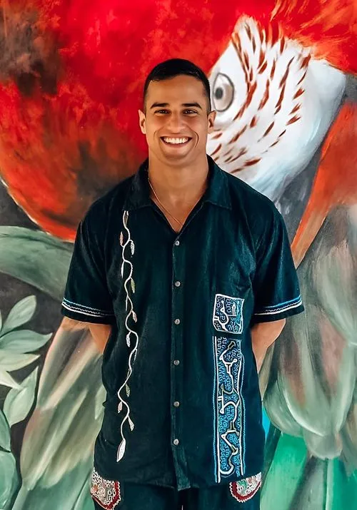 Smiling man wearing a traditional embroidered shirt stands in front of a vibrant jungle mural featuring a parrot, likely a guide or facilitator at a spiritual retreat.