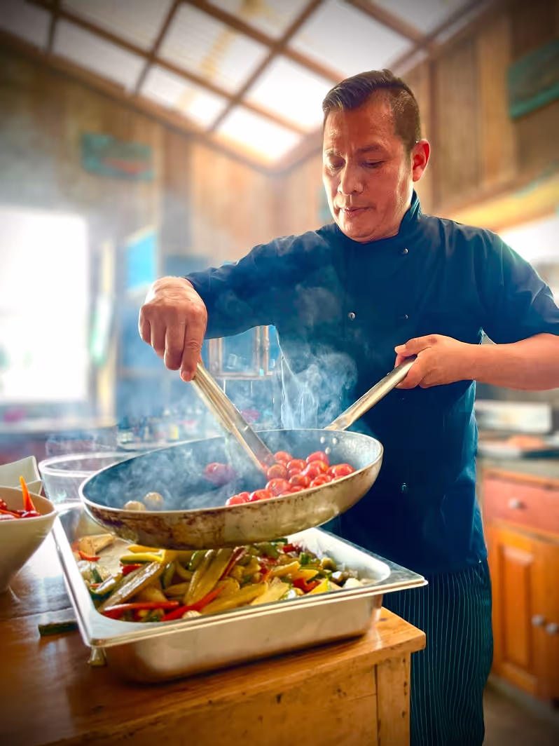 Chef plating sautéed vegetables from a steaming skillet into a tray inside a rustic wooden kitchen at Arkana spiritual retreat center.
