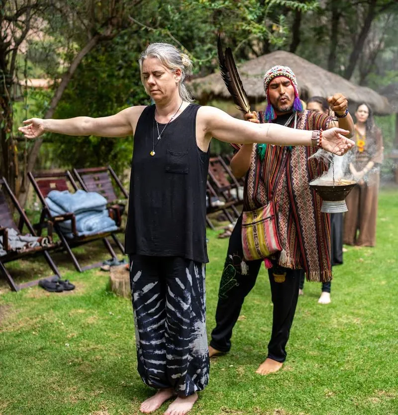 A woman stands barefoot with arms outstretched as a shaman performs a cleansing ritual using smoke and feathers in an outdoor setting, with others watching in the background.
