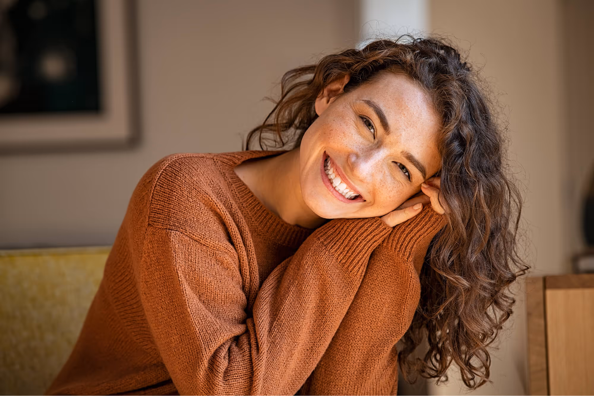A smiling woman in a brown sweater sitting on a couch.