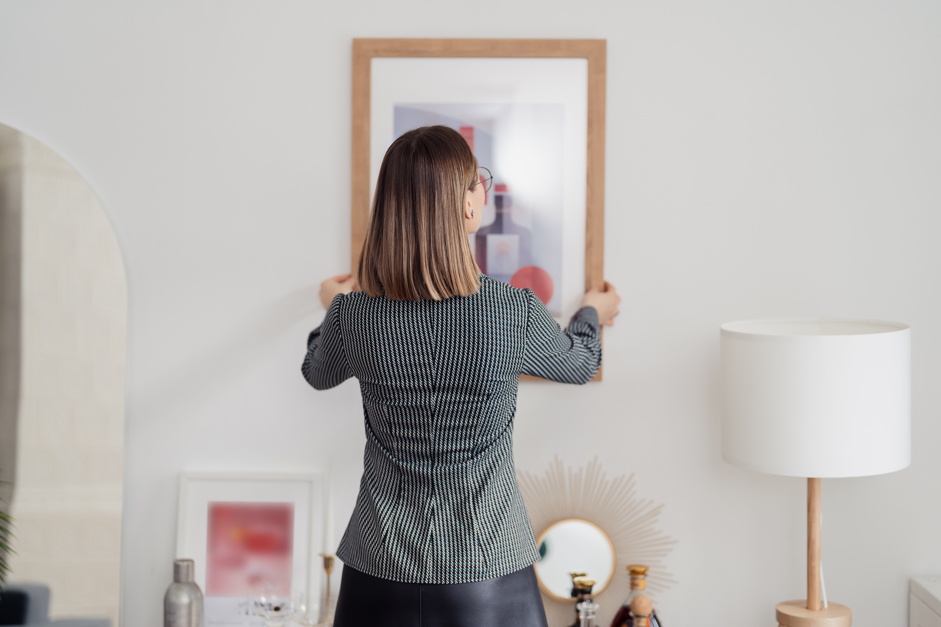 A woman hanging a framed picture on a white apartment wall, representing the ease of personalizing the spacious homes at Avian Apartments in Colorado Springs, CO.