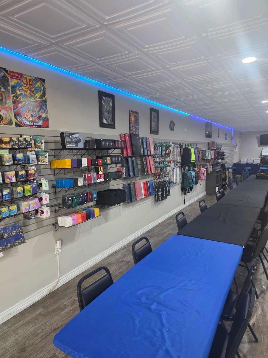 Game store interior with playmats, card sleeves, backpacks on wall and tables with black and blue covers.