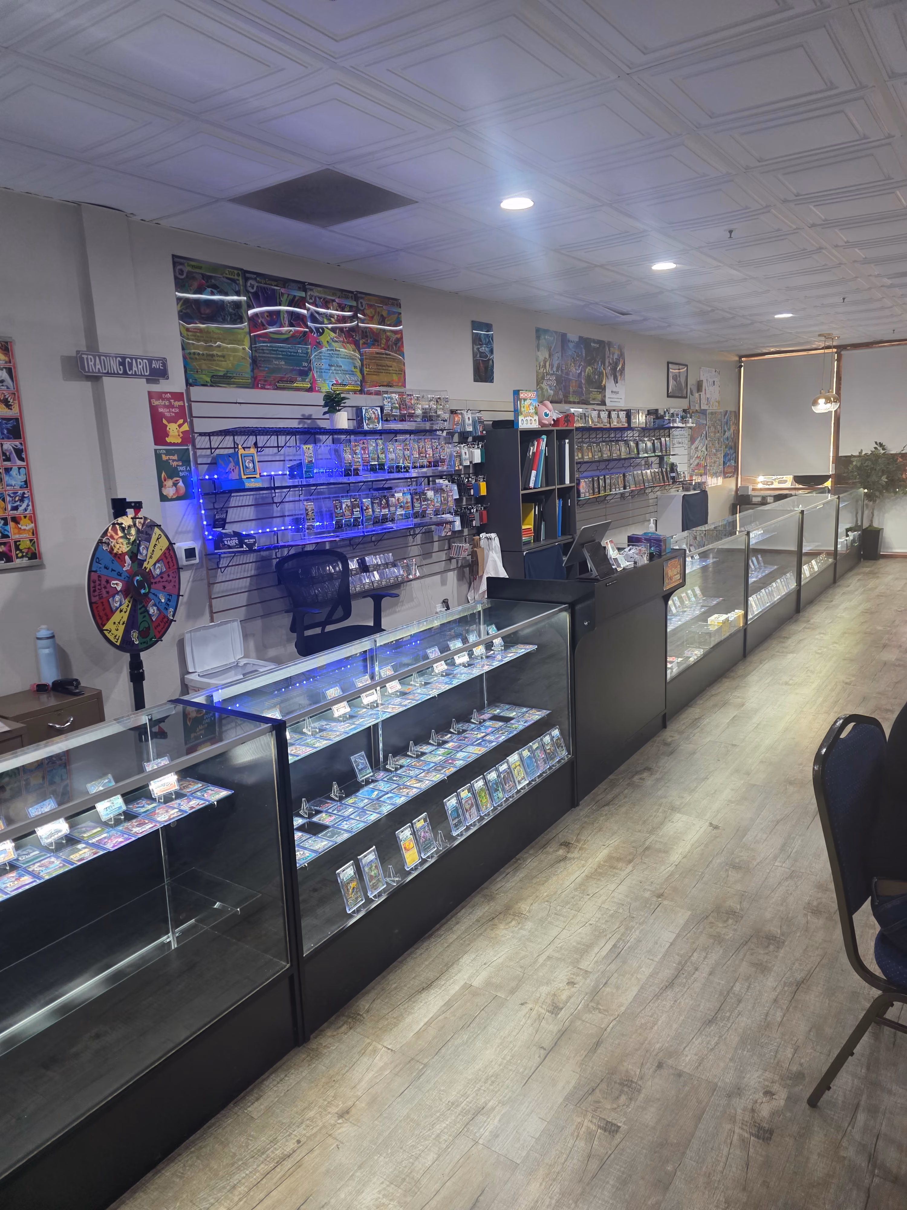 Interior of a trading card store with glass display cases filled with collectible cards, a spinning prize wheel, wall-mounted card racks, posters, and chairs along a wood floor.