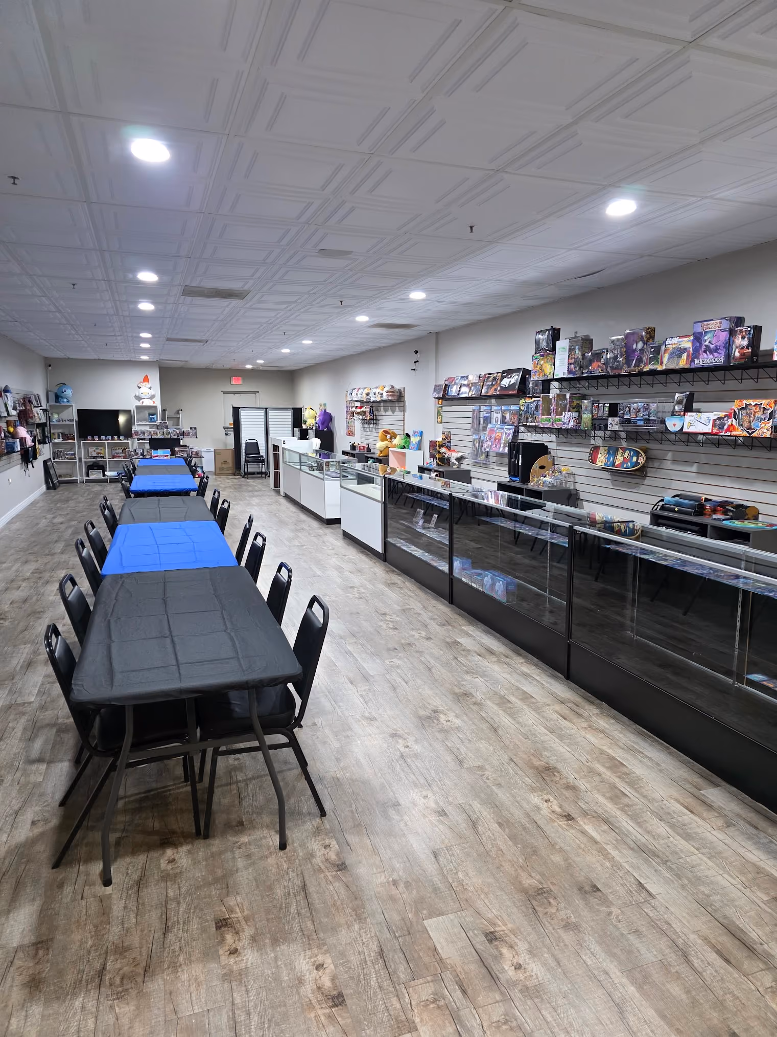 A spacious game store interior with wooden floor, long tables covered with black and blue tablecloths surrounded by chairs, and display shelves against the walls filled with various board games, card games, and collectibles.