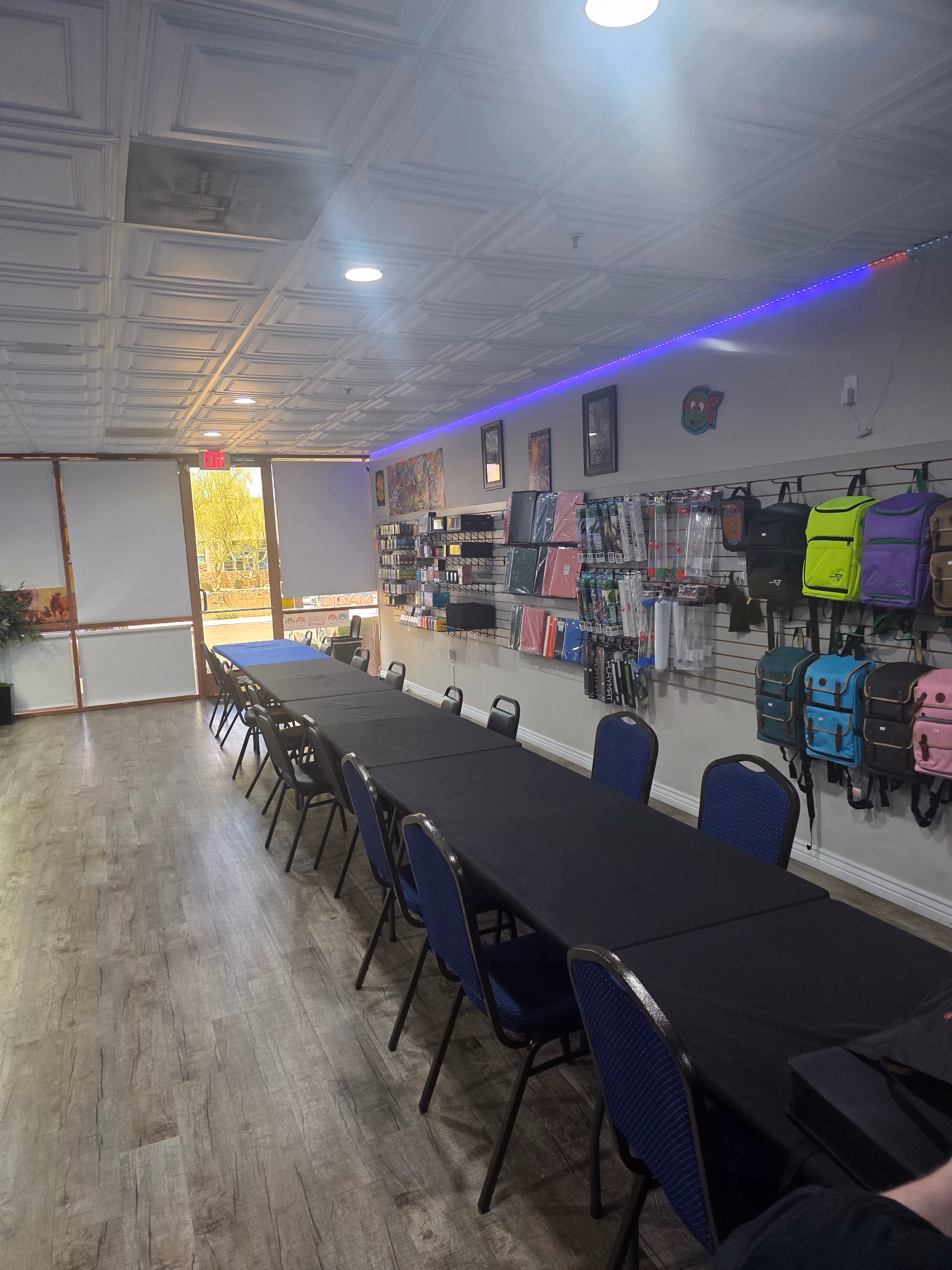 Interior of a game or hobby store with a long table covered in black tablecloths surrounded by chairs along the center. The right wall displays colorful backpacks and various gaming supplies hung on slat walls. The ceiling has recessed lighting and a strip of purple LED lights near the wall. The back features a glass door with an exit sign and windows letting natural light in.