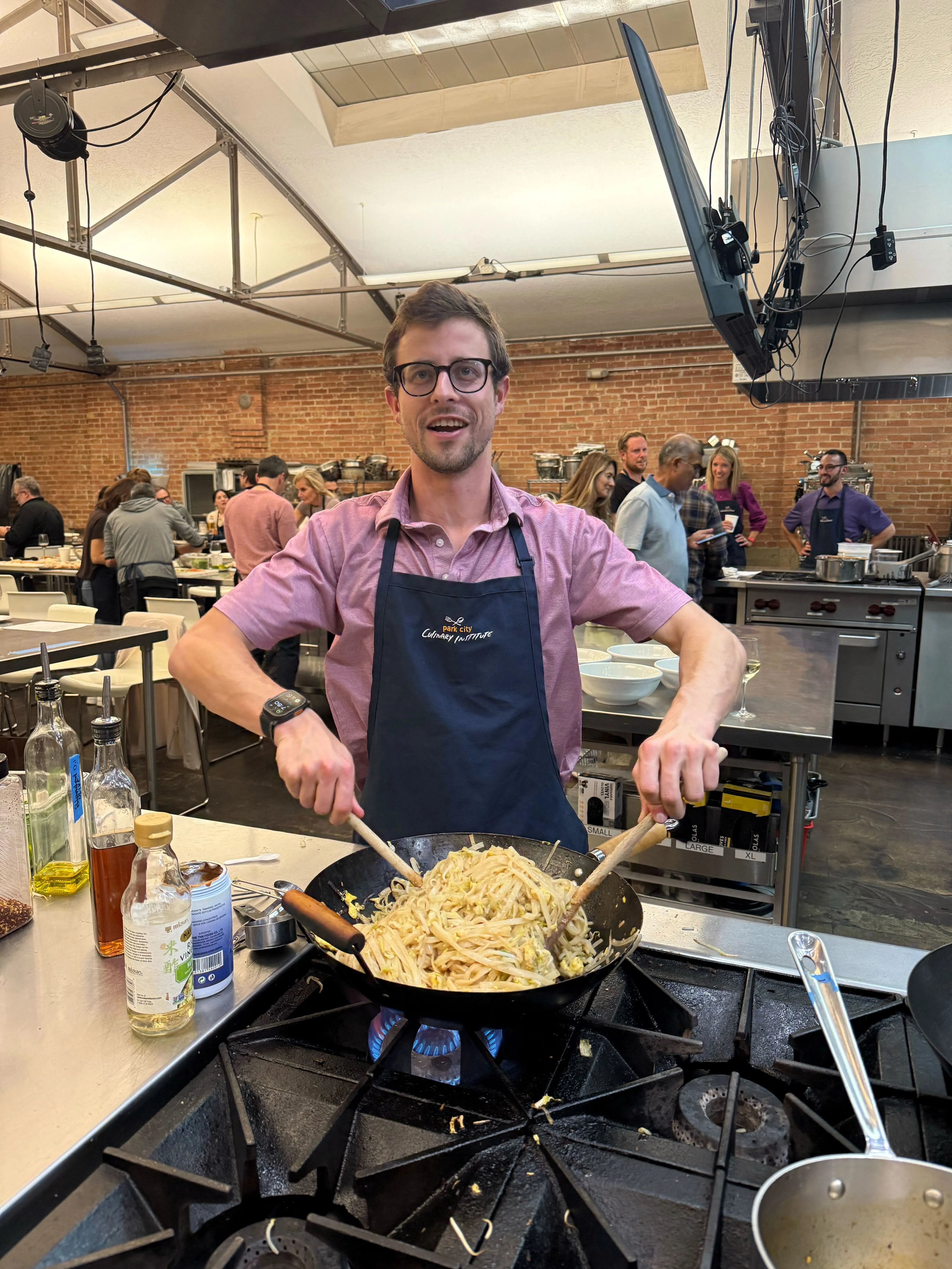 Man wearing glasses and a black apron cooking stir-fried noodles in a wok on a gas stove in a busy commercial kitchen.