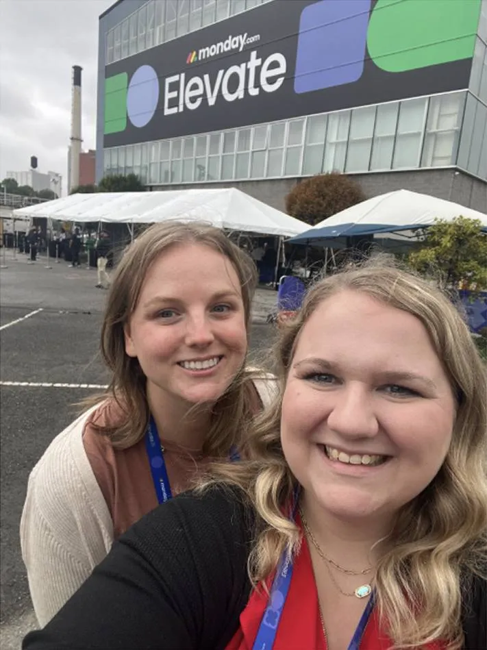 Two smiling women wearing lanyards taking a selfie outdoors in front of a building with a large banner that reads 'monday.com Elevate'.