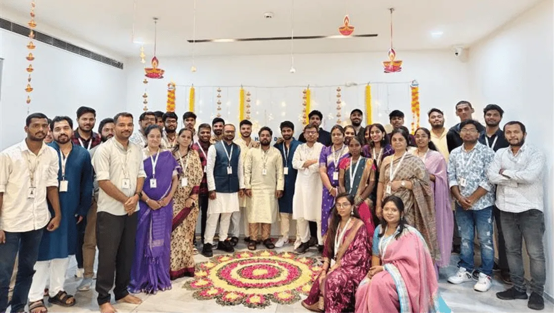 Group of men and women wearing traditional Indian attire standing and sitting around a colorful flower rangoli decoration indoors.