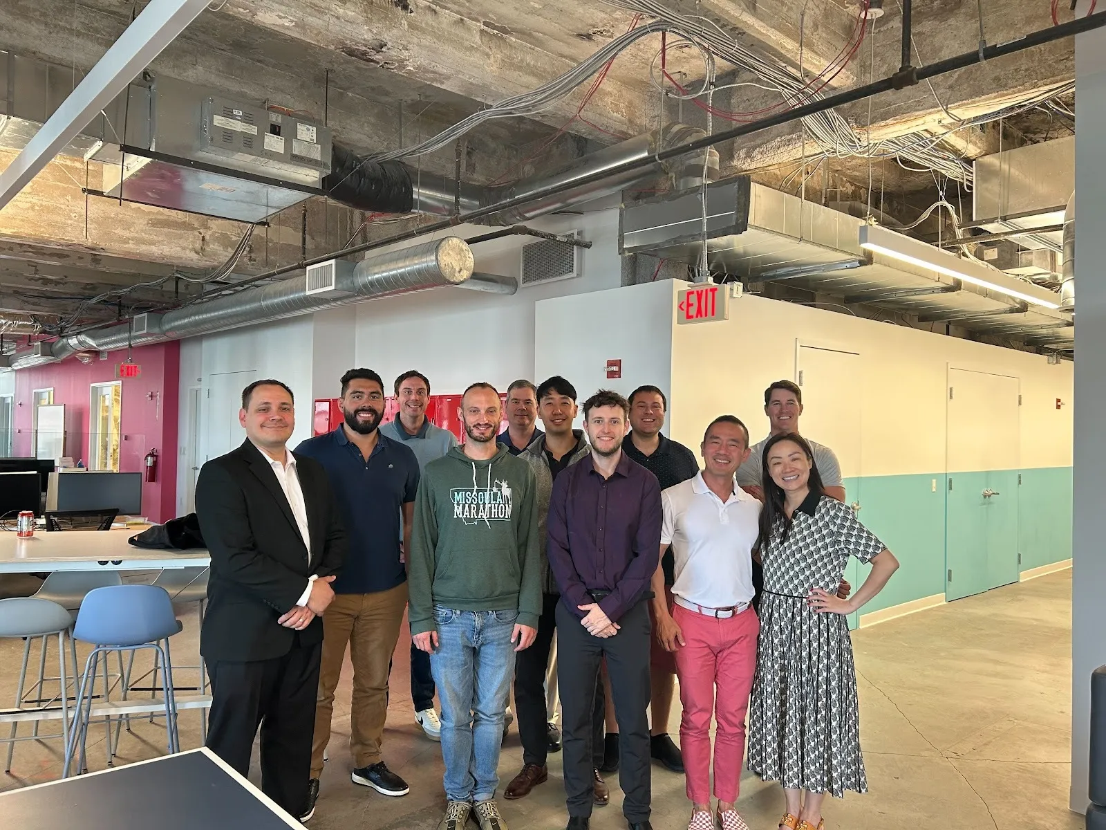 Group of ten people standing together and smiling in an industrial-style office with exposed ceiling pipes and ventilation.