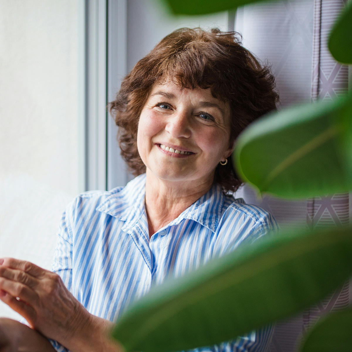 Smiling mature woman in a striped shirt sitting by a window surrounded by houseplants.