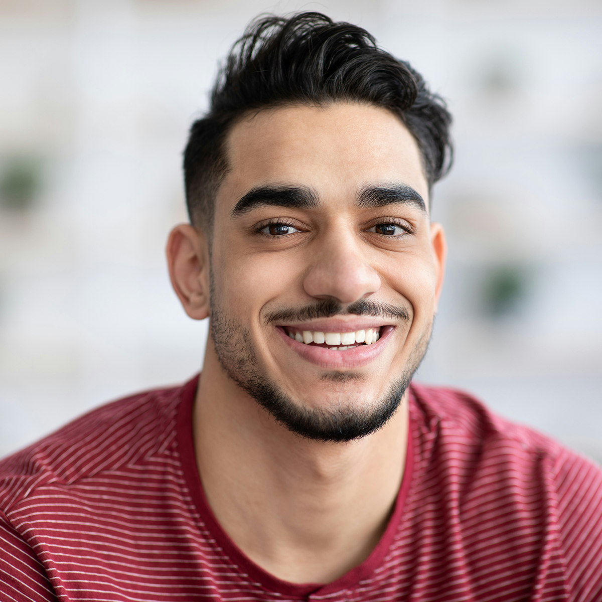 Cheerful young man in a red striped shirt smiling at the camera in a well-lit indoor setting.