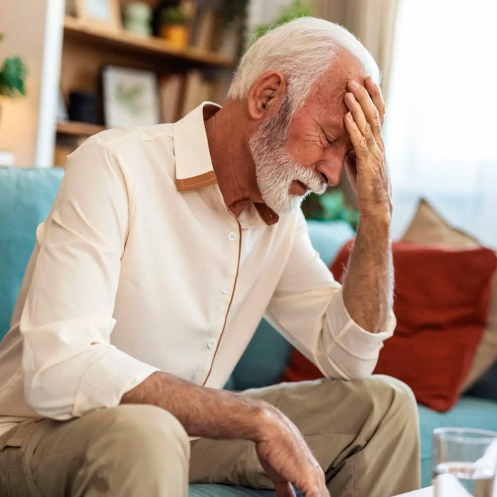 Senior man sitting on couch with hand on forehead, showing signs of brain fog or confusion.