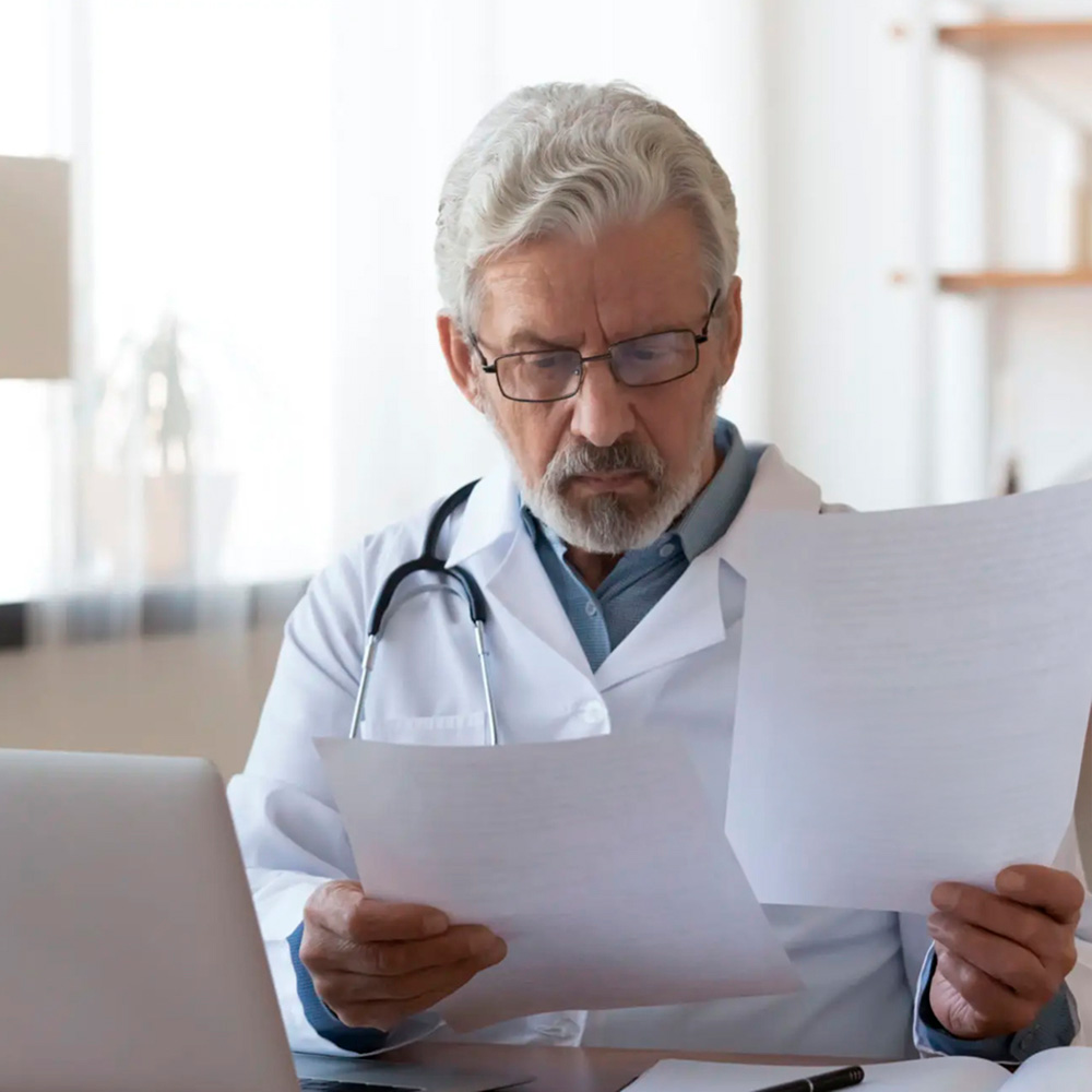 Senior male doctor reviewing medical documents at his desk with a laptop.