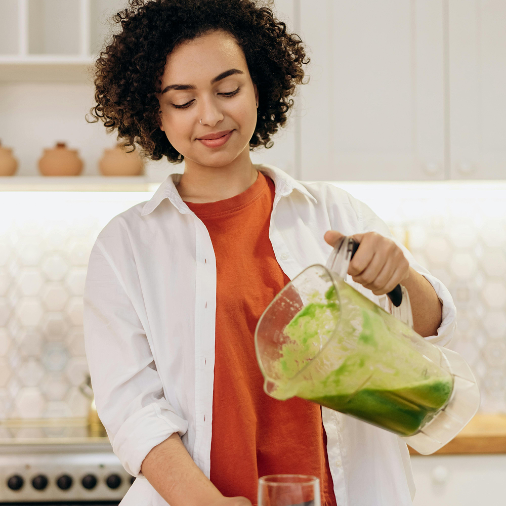 Smiling woman in a kitchen pouring a green smoothie from a blender into a glass, suggesting focus on healthy eating and nutrition.