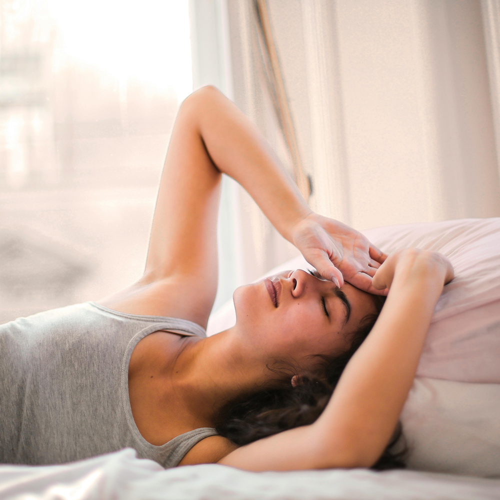 Young woman lying in bed with a pained expression, holding her forehead, possibly experiencing menstrual discomfort or fatigue.