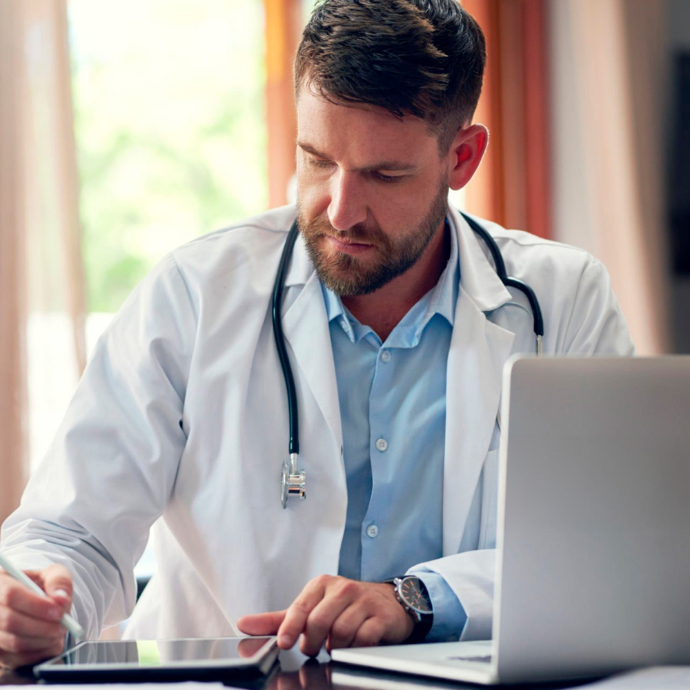 Male doctor in a white coat reviewing information on a tablet while seated at a desk with a laptop, appearing focused and professional.