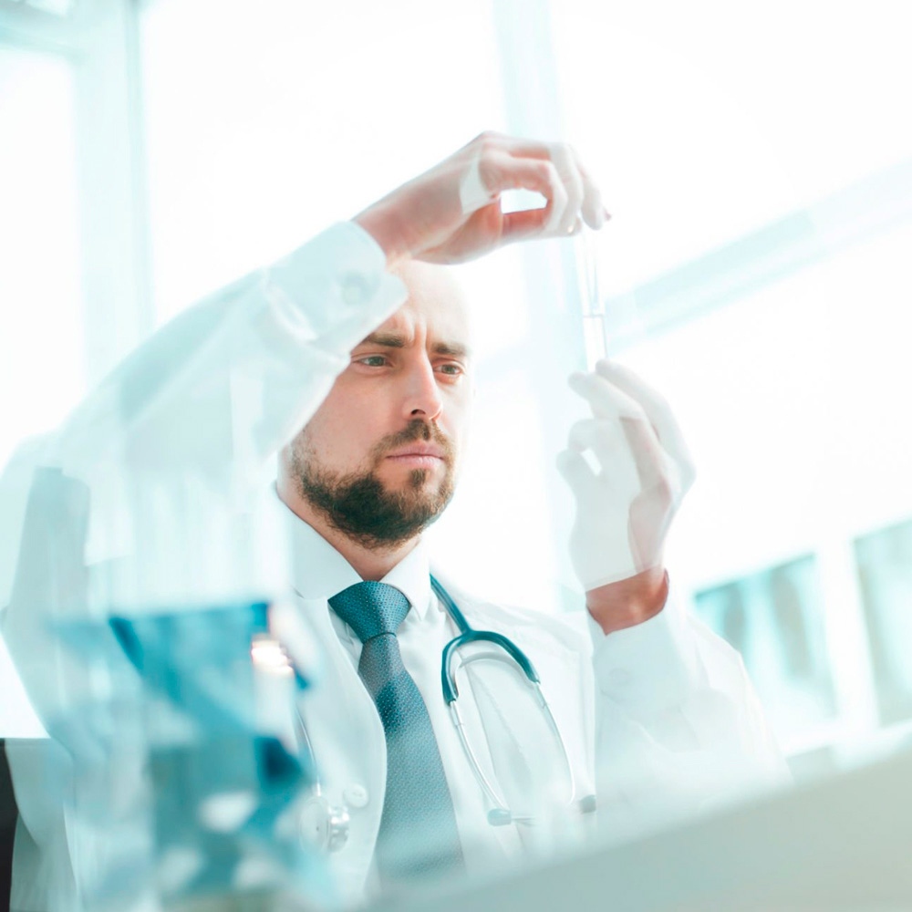 Focused male doctor in a lab coat and stethoscope examining a test tube in a bright clinical setting.