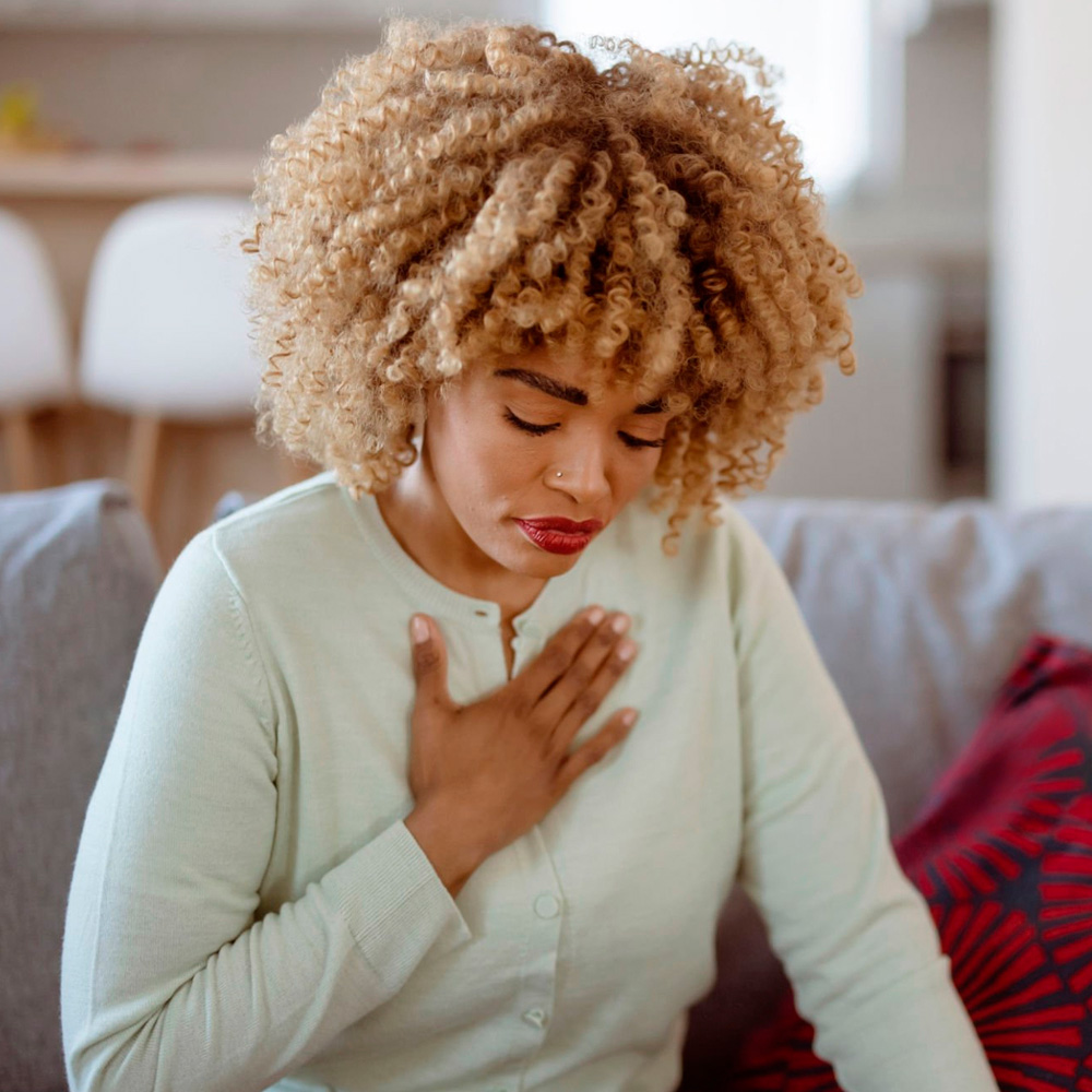 Woman sitting on a couch, holding her chest in discomfort, showing signs of chest pain or heart-related symptoms.
