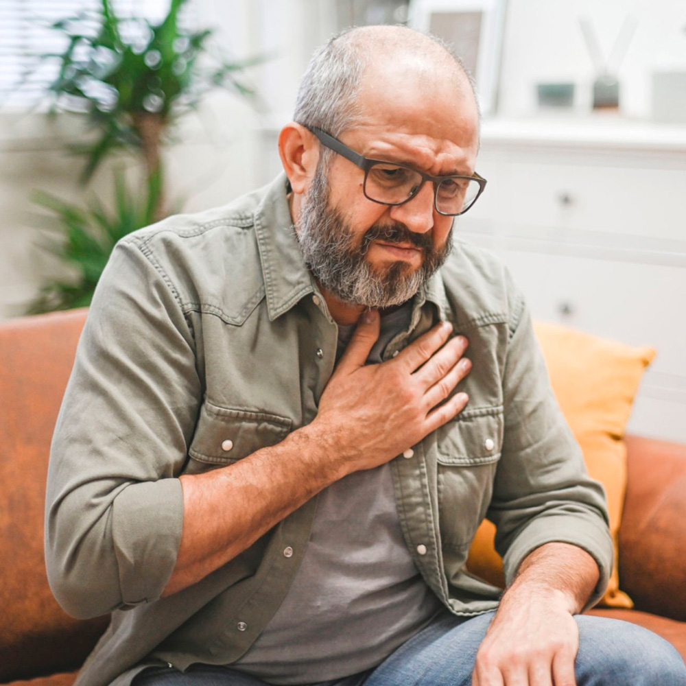 Middle-aged man sitting on a couch, holding his chest and appearing distressed, possibly experiencing shortness of breath or breathing difficulties.