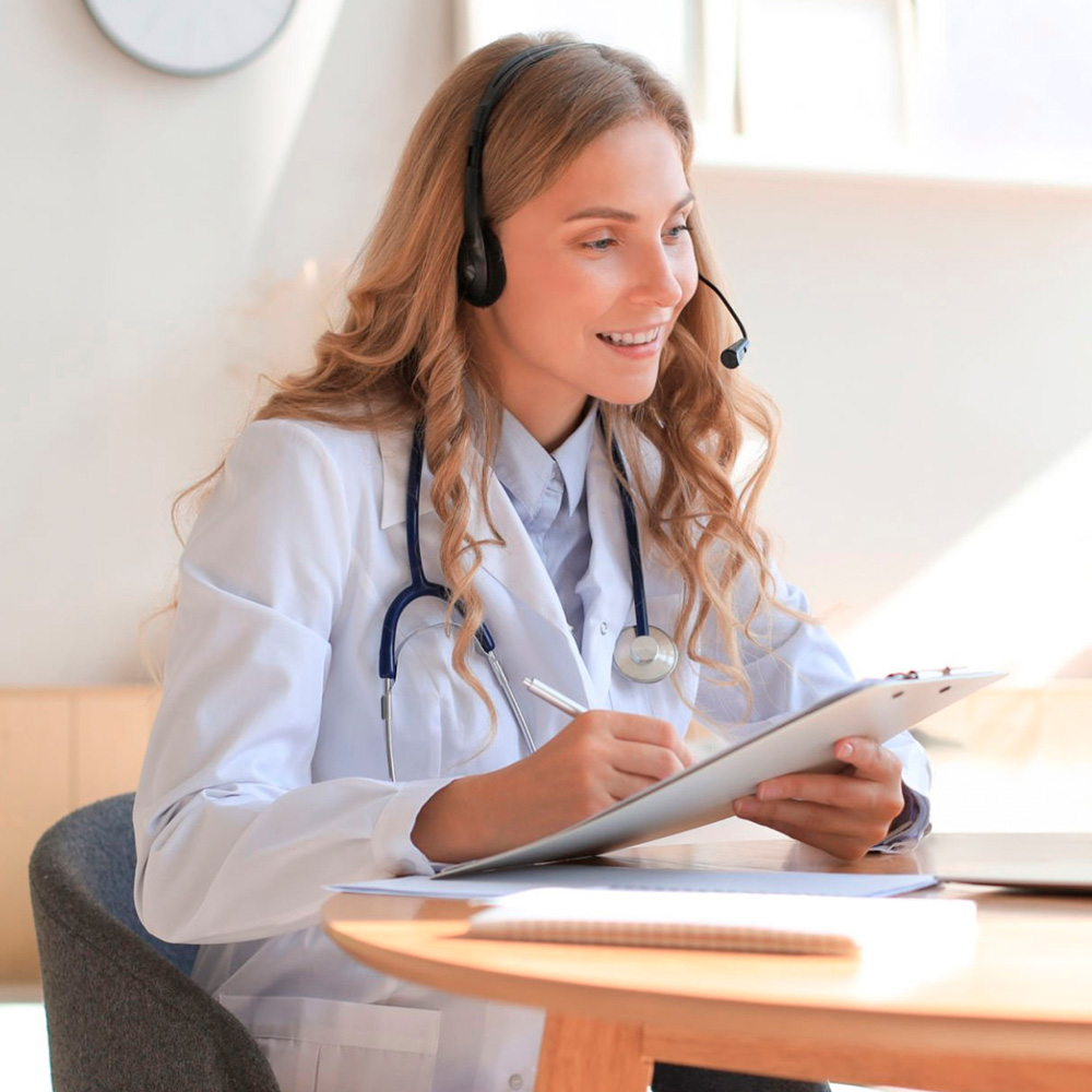 Female doctor with headset writing notes during telehealth consultation.