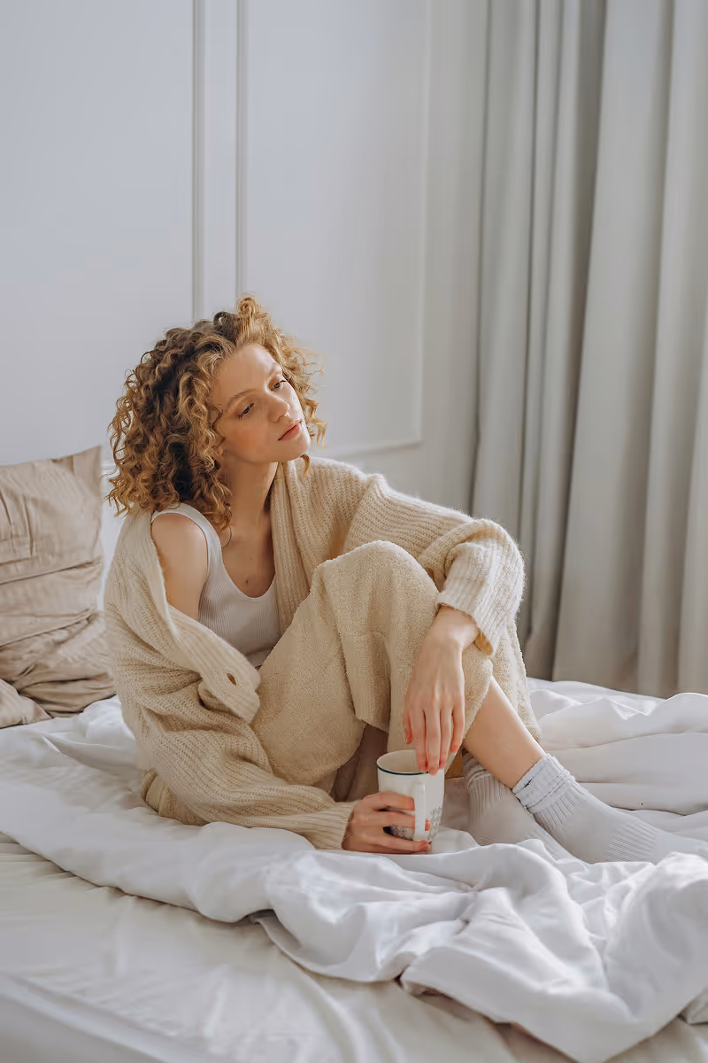 Woman with curly hair in beige cardigan sitting on bed with a cup, Hedfirst PCOS treatment lifestyle photo.