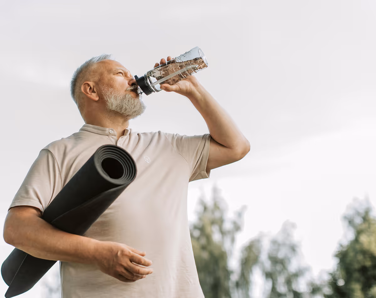 Older man with yoga mat drinking water.
