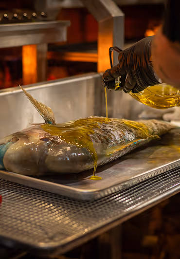A whole fish on a metal tray is being drizzled with olive oil by a person wearing black gloves. The scene conveys a sense of culinary preparation.