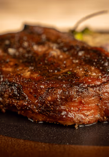 Close-up of a juicy, grilled steak on a dark surface, seasoned with coarse salt and black pepper. The rich, brown crust suggests a savory, flavorful meal.