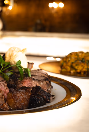 A close-up of a gourmet meal featuring sliced roast beef garnished with parsley and a dollop of sauce. A vegetable side dish is blurred in the background. The setting is elegant, with warm lighting creating a luxurious ambiance.