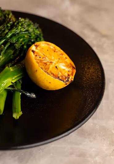 Grilled lemon half next to green broccolini on a black plate. The setting is elegant with a rustic feel, emphasizing a fresh, flavorful meal.