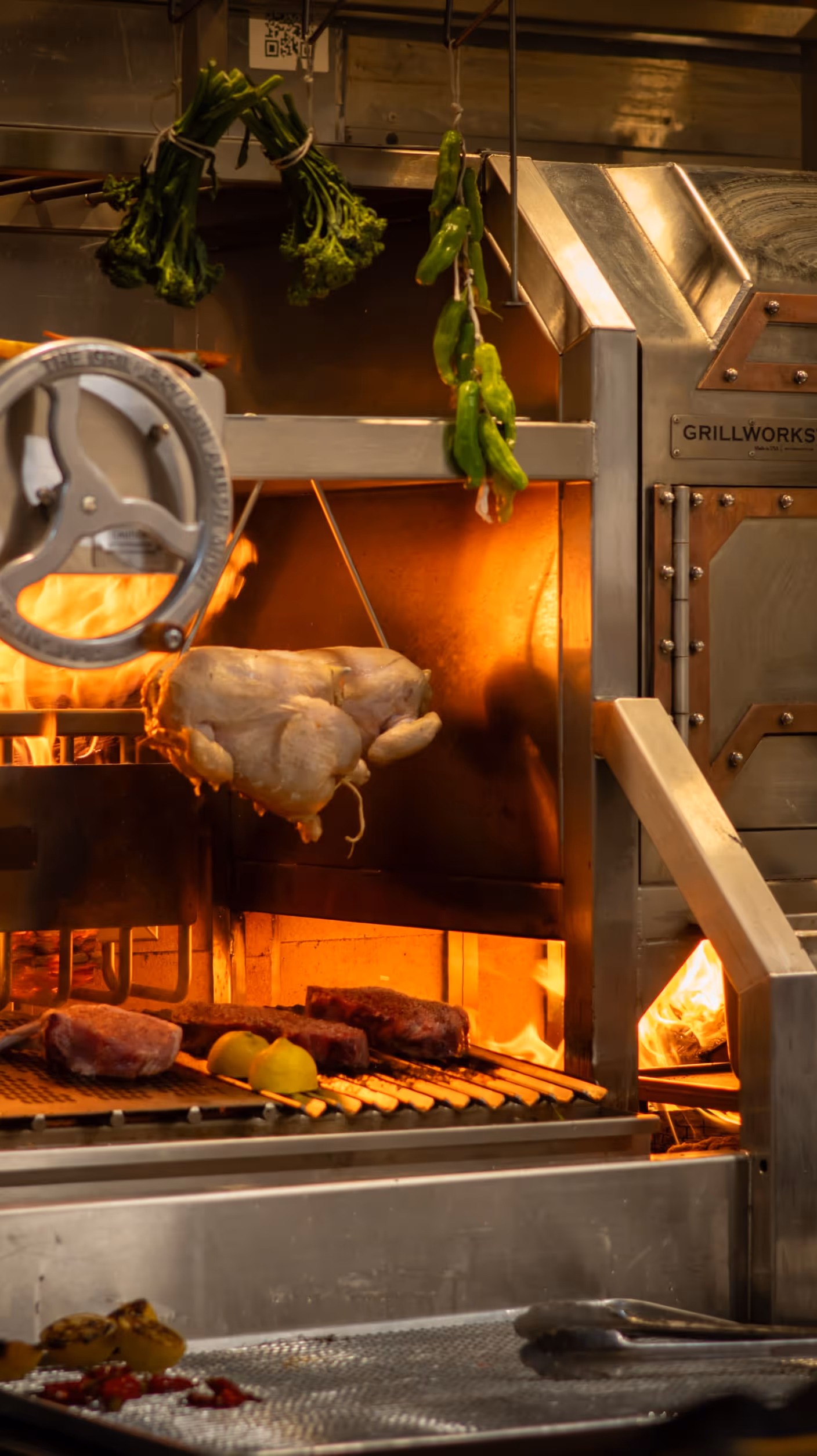 Commercial kitchen scene with a rotisserie oven. A chicken hangs above a grill with flames, surrounded by vegetables and meat. The ambiance is warm and busy.