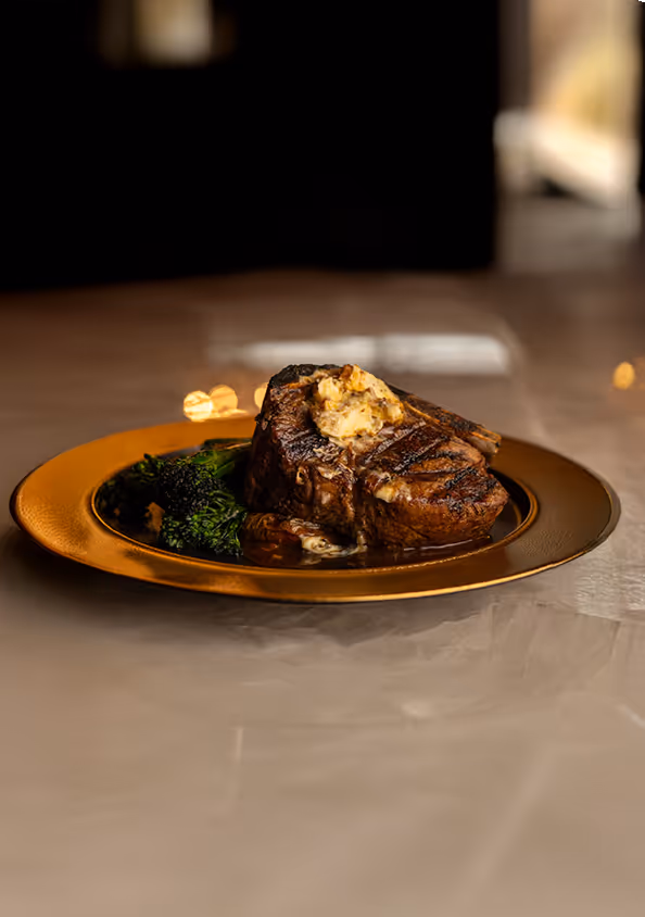 Grilled steak with herb butter on a bronze plate, accompanied by bright green broccolini. The setting is warm, evoking a luxurious dining experience.