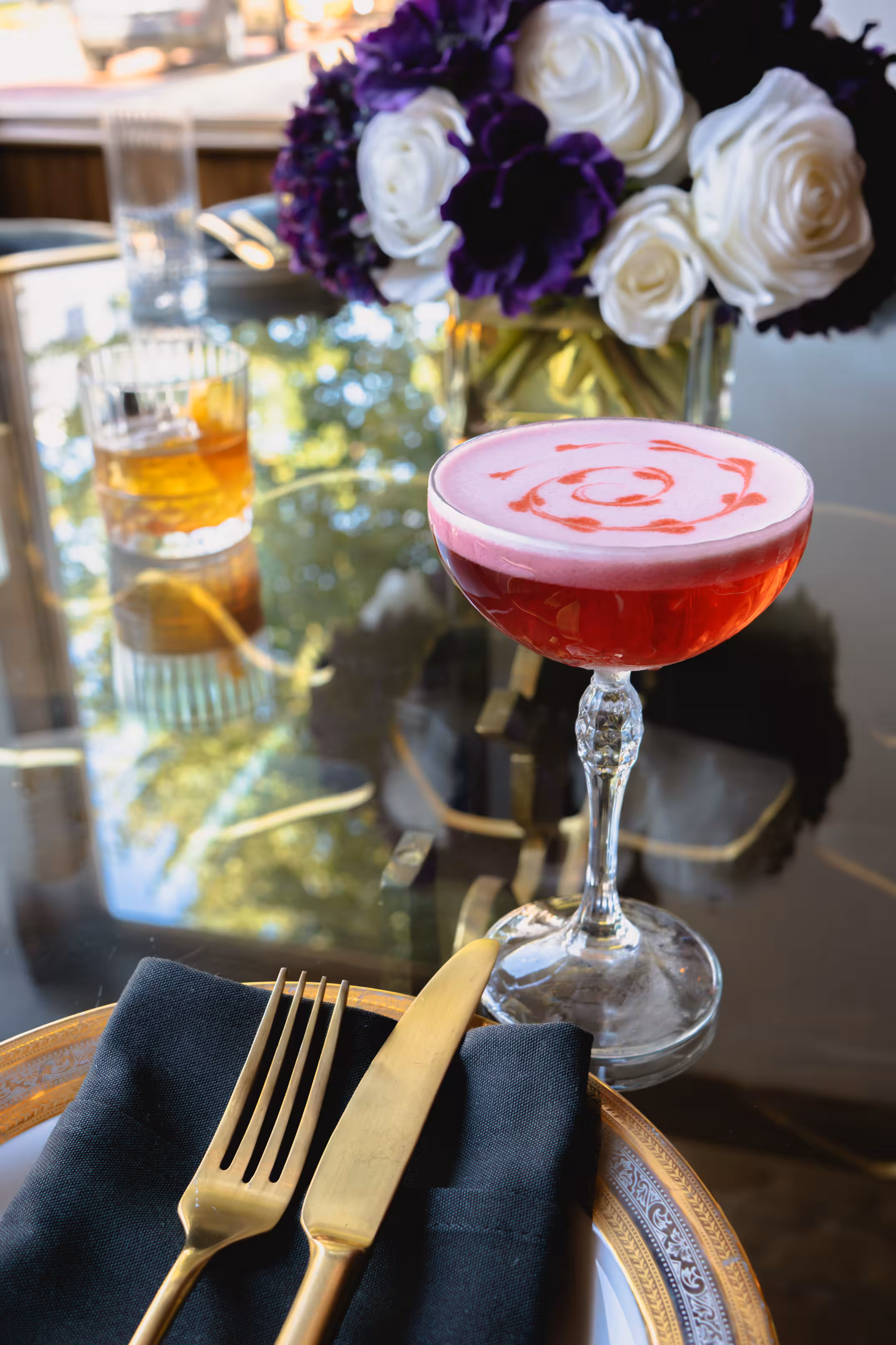 A pink cocktail with frothy topping and swirl design sits on a reflective table beside a whiskey glass, gold cutlery on a napkin, and a rose bouquet.