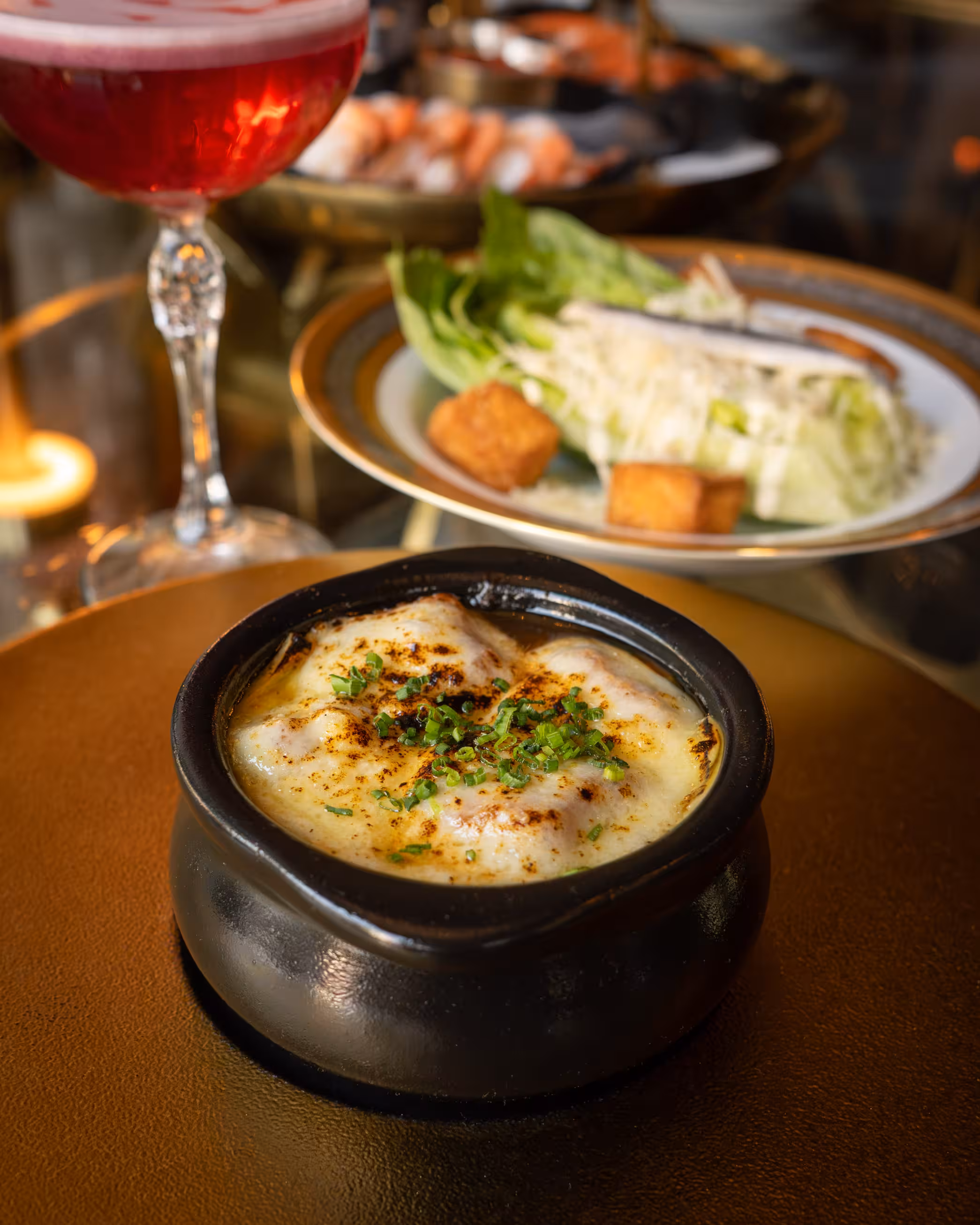 Crème brûlée in a black ramekin topped with chives, set on a brown plate. A vibrant pink cocktail and a lettuce dish are blurred in the background.